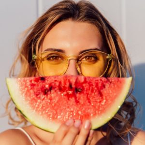 Charming lovely woman in yellow sunglasses hiding a half of her face with piece of watermelon, joking, standing against the wall, outdoors.