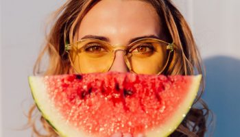 Charming lovely woman in yellow sunglasses hiding a half of her face with piece of watermelon, joking, standing against the wall, outdoors.